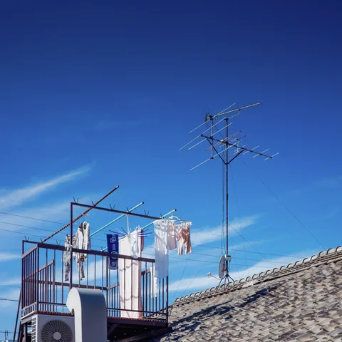 Laundry hangs from a rooftop railing beside a tall television antenna above gray tiles and blue sky.