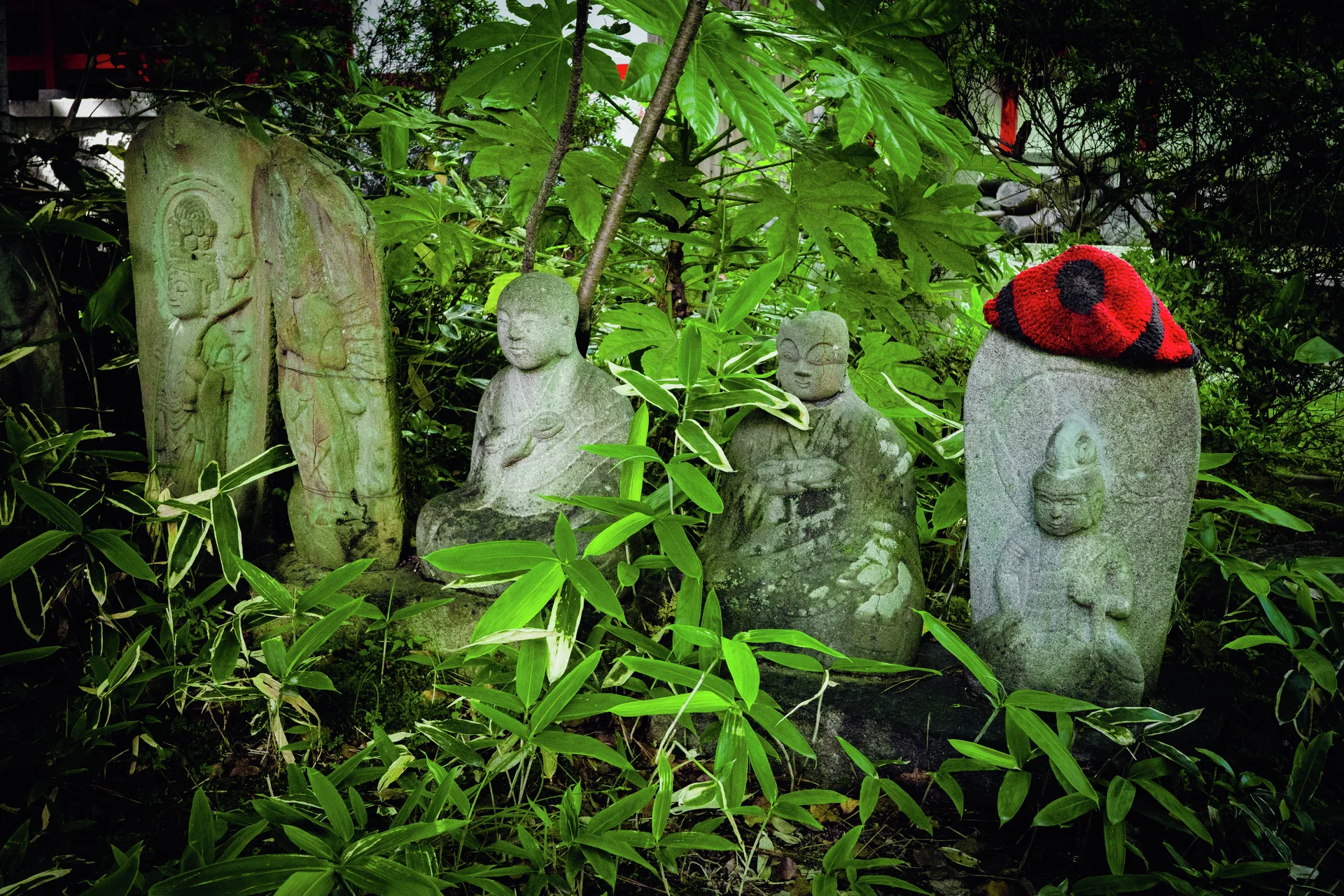 Several weathered stone figures stand among dense green foliage, with red fabric covering one rounded slab.