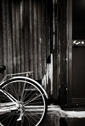A bicycle's rear wheel rests beside weathered vertical siding and a dark door in black and white.