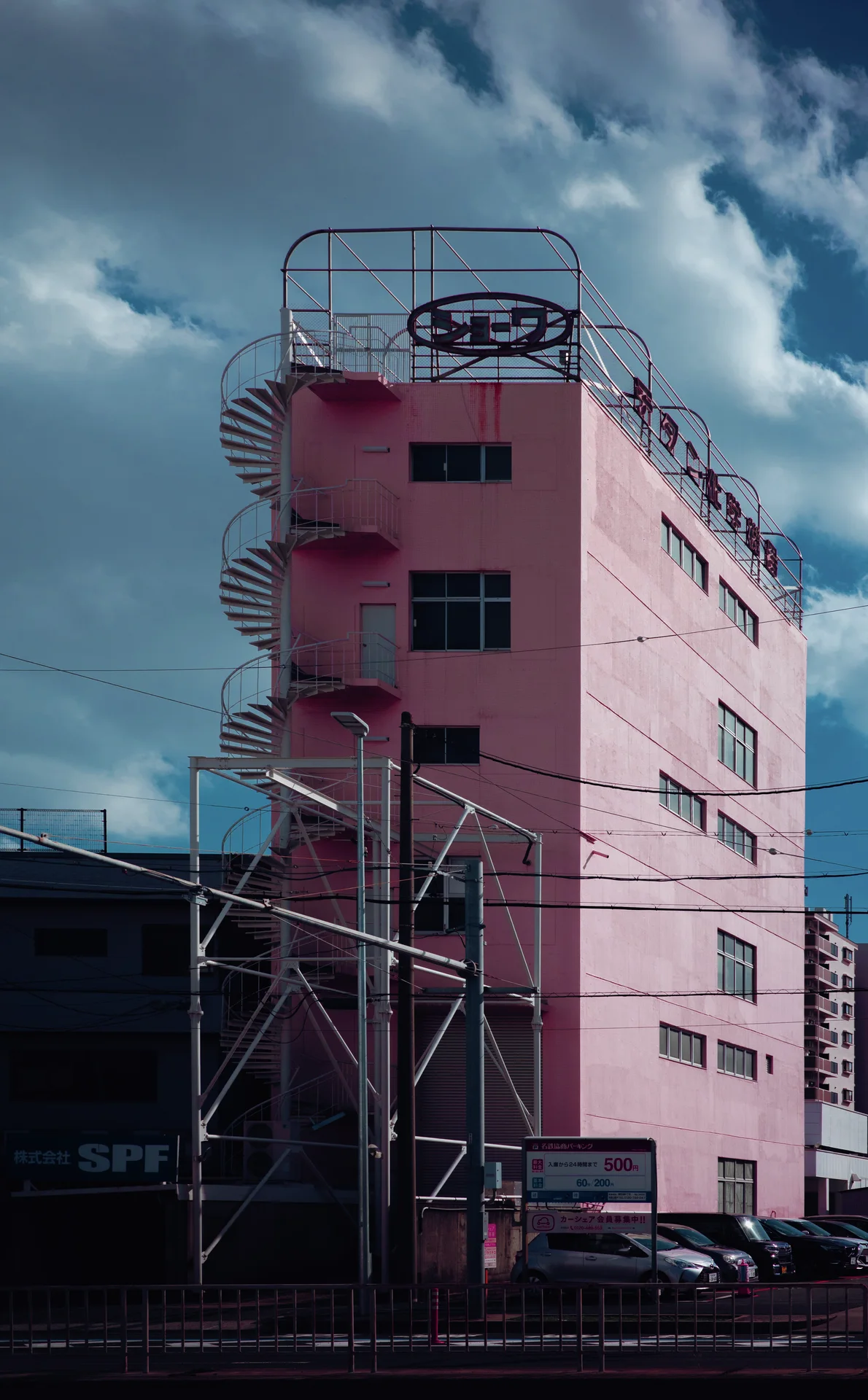 A pink multistory building has an external spiral staircase and rooftop metal framework beneath billowing clouds.
