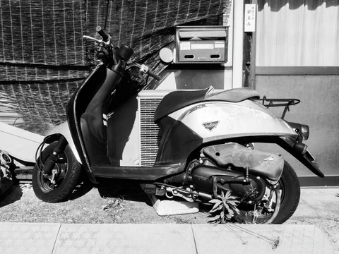 A worn scooter is parked beside a mailbox and outdoor air-conditioning unit in black and white.
