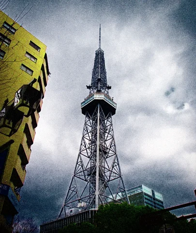 A tall steel lattice tower rises beneath gray clouds beside a yellow building and dark treetops.