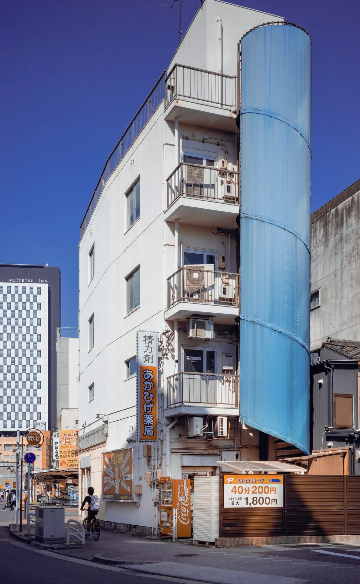 A narrow white apartment building has small balconies beside a tall curved blue exterior panel.