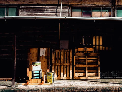 Wooden pallets and small boxes lean against a shadowed wooden building beneath old upper windows.