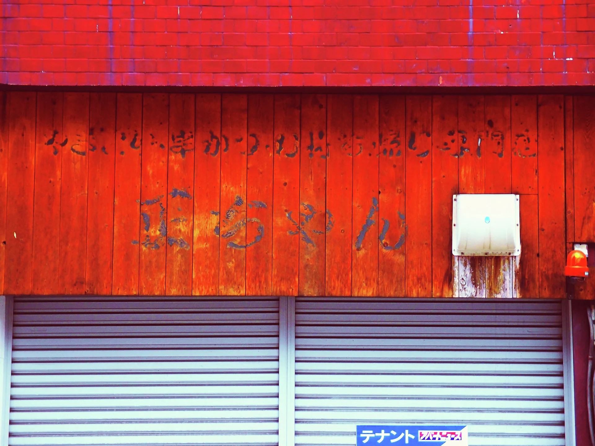 Faded lettering covers orange wooden panels above closed gray shutters and a stained wall fixture.