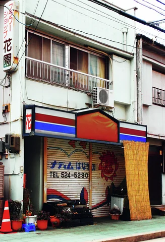 A closed narrow storefront has a striped sign, upper balcony, and colorful buckets beside its shutter.