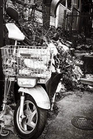 A white scooter with a wire basket stands among weeds beside a building in black and white.