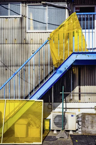 A blue exterior staircase has bright yellow mesh draped over its railings beside corrugated walls.