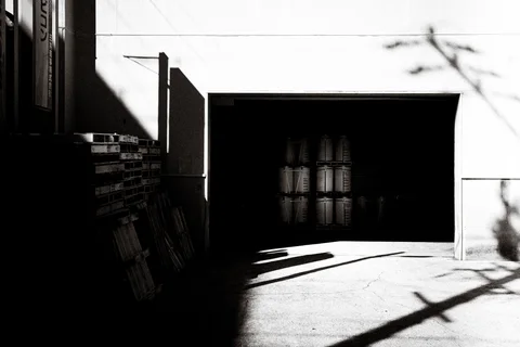 A sunlit industrial entrance reveals stacked cylinders inside darkness, crossed by strong diagonal shadows.