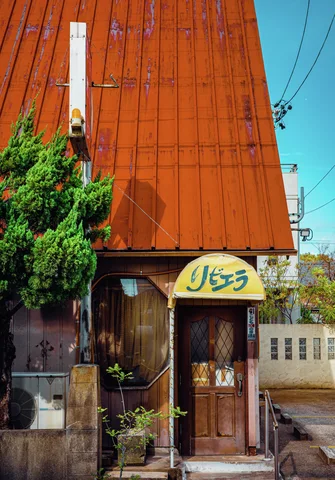 A steep orange metal facade towers above a narrow wooden entrance and a small yellow sign.