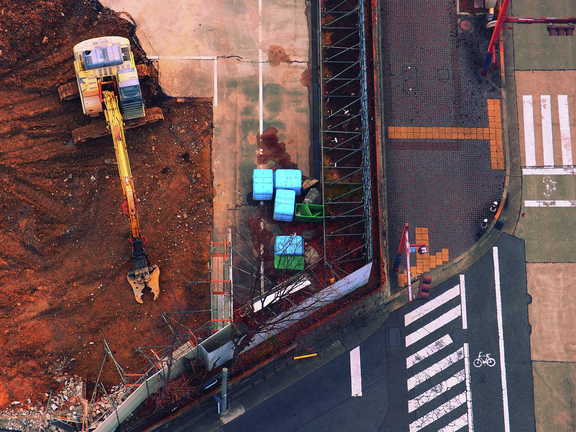 A yellow excavator stands beside a fenced roadside work area, blue containers and white crossing stripes.
