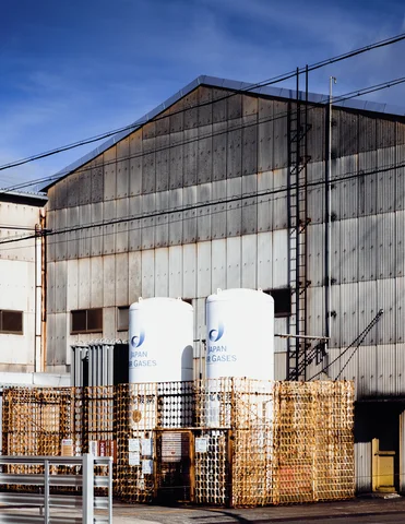 Two white cylindrical tanks stand inside a rusty fence in front of a large corrugated warehouse.