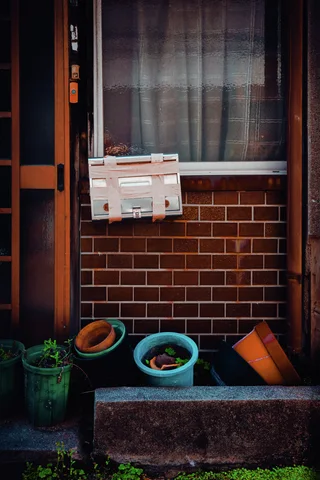 A pale mailbox hangs below a curtained window above colorful pots on a brick-fronted wall.