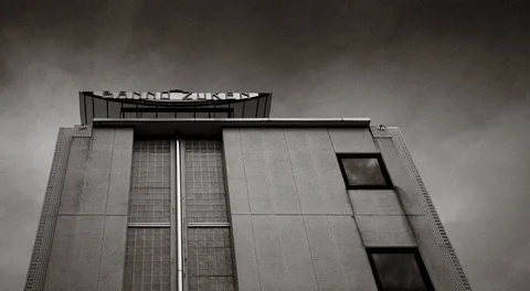 A plain building rises beneath dark clouds, with narrow vertical windows and rooftop framework in black and white.