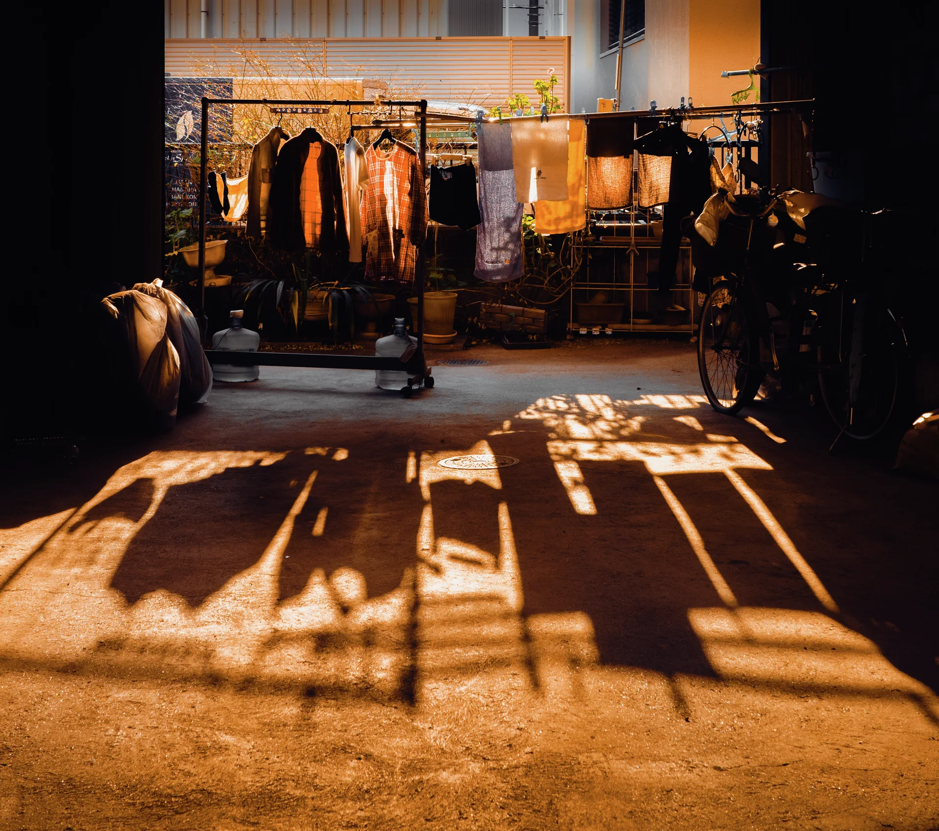 Warm sunlight casts long clothing-rack shadows across a passage leading to hanging garments and bicycles.