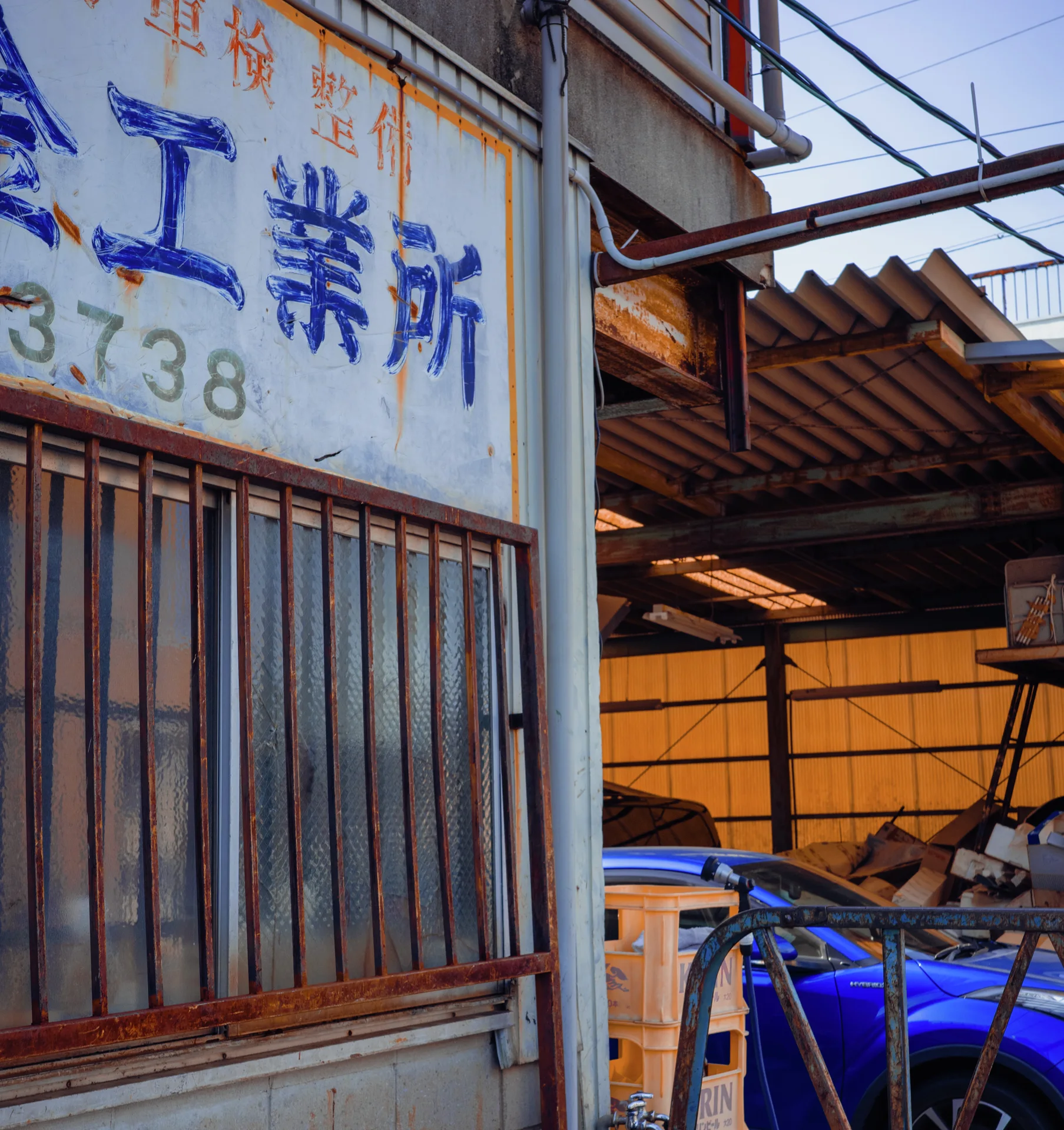 A faded Japanese workshop sign sits above barred windows beside an open shed containing blue equipment.