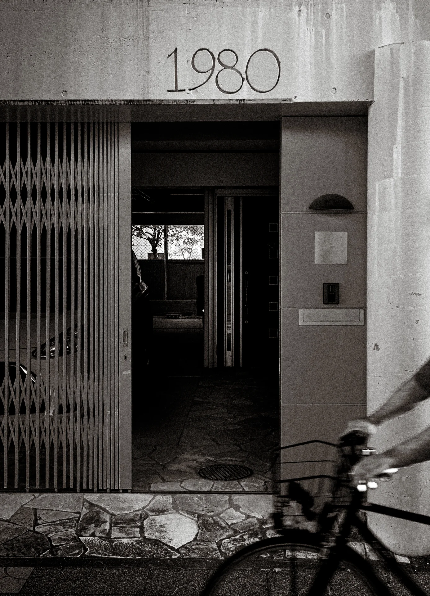 The number 1980 appears above a shadowy entrance as a cyclist passes in black and white.