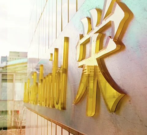 Thick gold lettering projects from a pale wall and casts sharp shadows in angled sunlight.