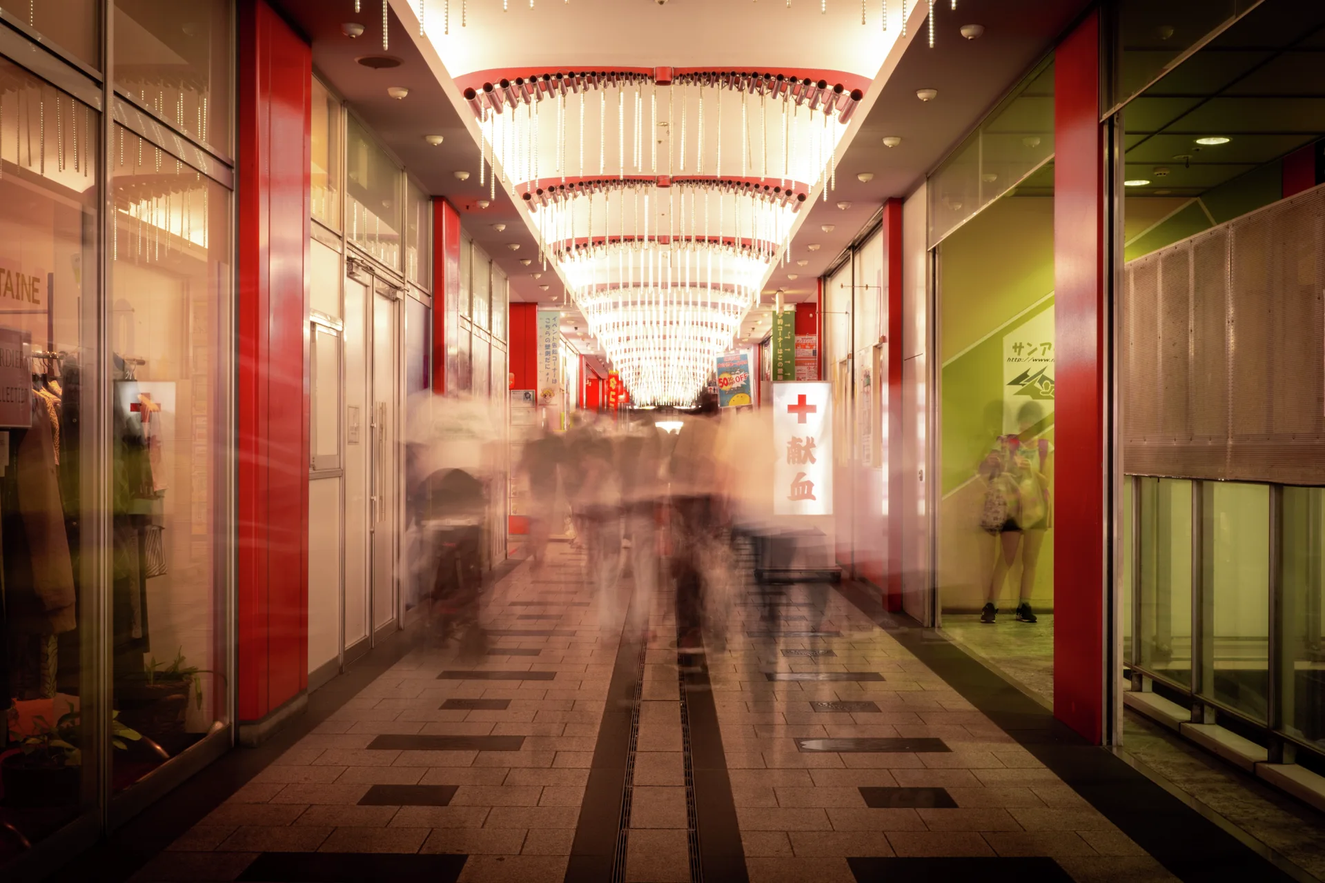 Blurred pedestrians pass through a brightly lit corridor lined with glass storefronts and red pillars.