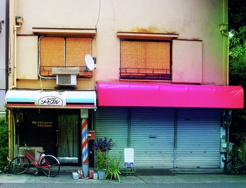 A striped barber pole stands beside closed blue shutters beneath a bright pink awning and bamboo blinds.