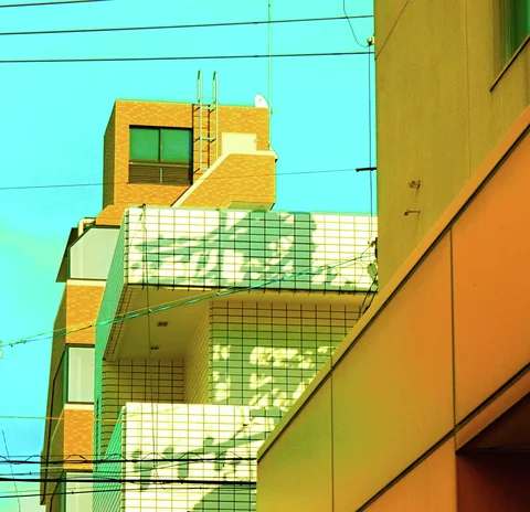 An exterior staircase casts angular shadows across a tiled building beside an orange wall under turquoise sky.