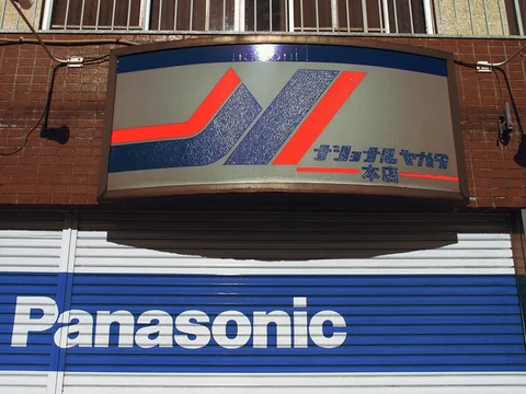 A curved geometric sign hangs above blue shutters bearing large white Panasonic lettering.