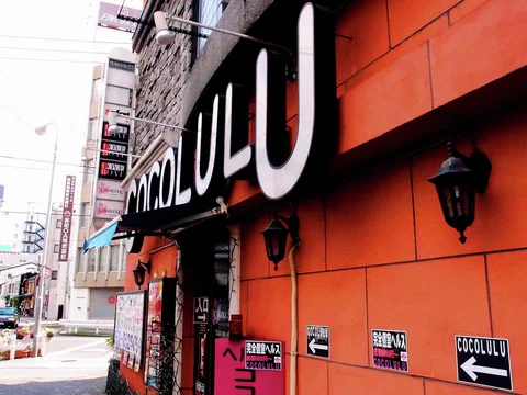 Large pale letters project above an orange storefront wall with dark lamps and directional notices.
