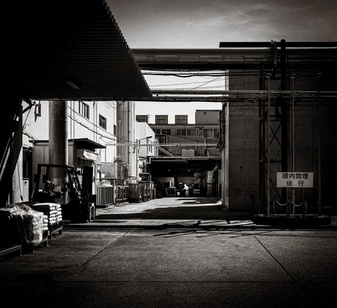 An industrial lane runs beneath overhead pipes between shadowed buildings in black and white.