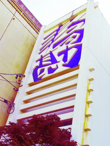 Large purple characters climb a tall pale facade above repeated horizontal ledges and dark foliage.