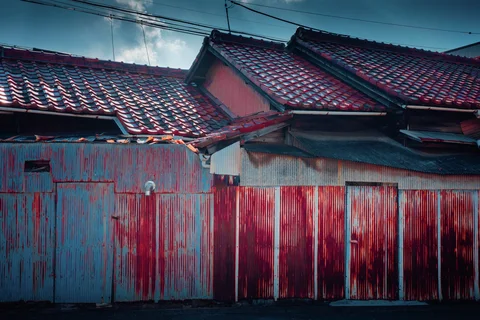 Red rust streaks cover blue and pale corrugated panels beneath layered dark tiled roofs.