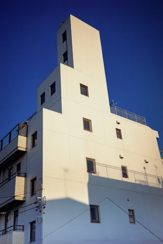 A tall narrow white building catches sunlight above deep shadows against a dark blue sky.
