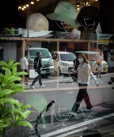 Pedestrians and parked vehicles reflect across a shop window displaying pale hats above leafy plants.