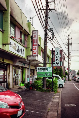 A red car occupies the foreground of a shop-lined street crowded with poles and overhead wires.