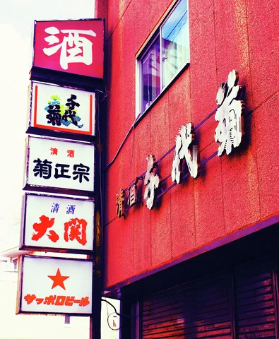 Stacked square drink-brand signs stand beside pale raised Japanese lettering on a red storefront.