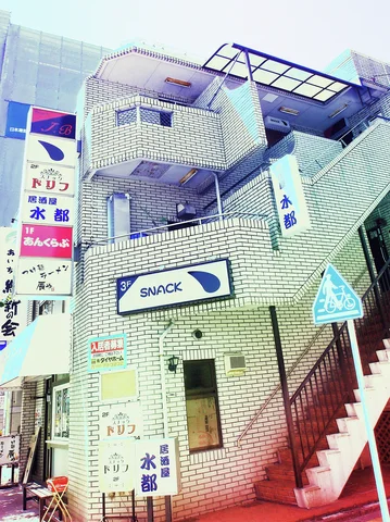 Exterior stairs climb a pale tiled building crowded with narrow shop signs and projecting balcony shapes.