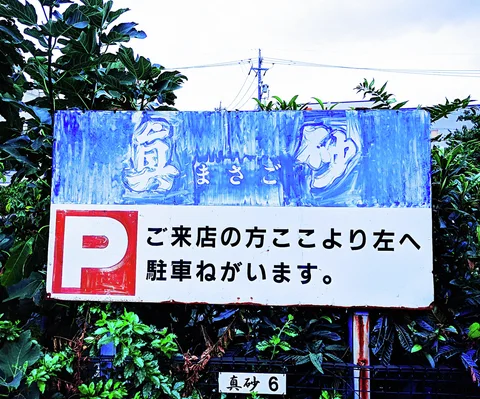A weathered blue and white parking sign stands among dense shrubs with bold red and black markings.