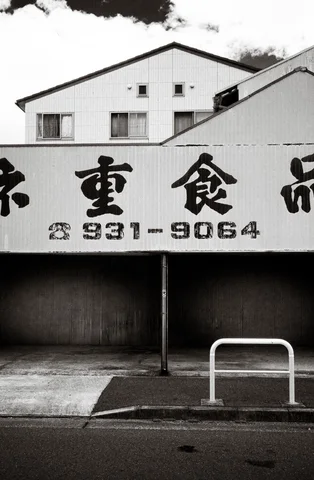 Bold Japanese lettering and telephone numbers span a low storefront sign above a roadside barrier in black and white.