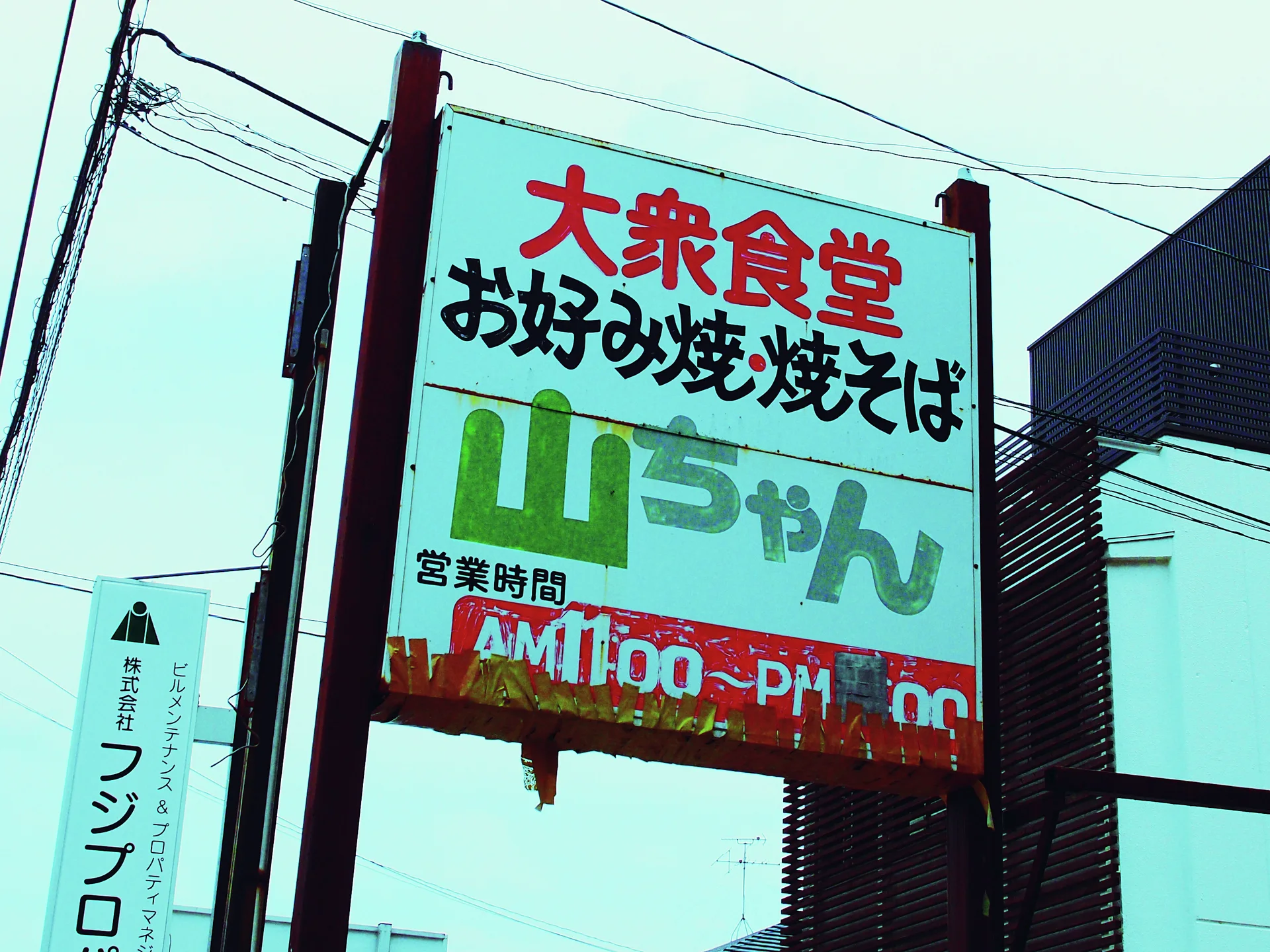 A weathered tall sign carries red, black, and green Japanese lettering beneath overhead wires.