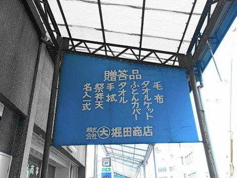 A blue Japanese shop sign hangs beneath a translucent covered walkway supported by dark metal beams.