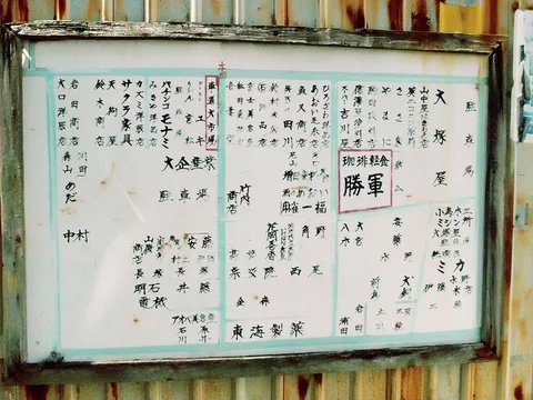 Columns of small handwritten Japanese characters fill a weathered framed board against rust-streaked corrugated siding.