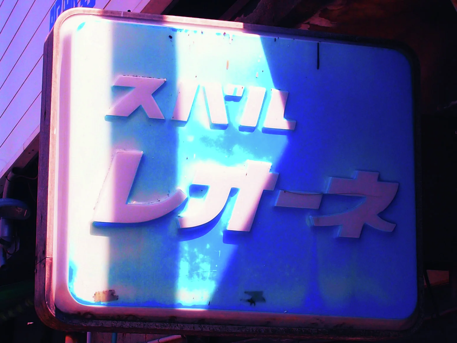 A shaft of sunlight illuminates raised pink Japanese lettering on a rounded blue projecting sign.
