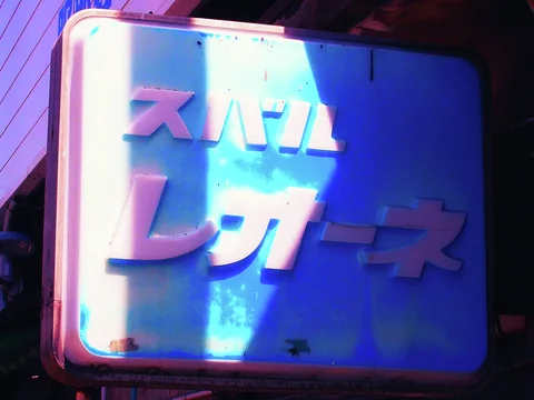 A shaft of sunlight illuminates raised pink Japanese lettering on a rounded blue projecting sign.
