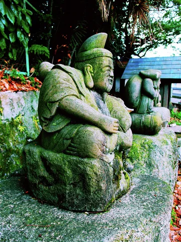 A seated weathered stone figure wearing a cap holds a fishing rod beside a small stone lantern.