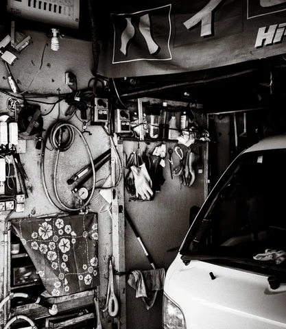 Hoses and tools hang densely inside a cluttered workshop beside a white vehicle in black and white.