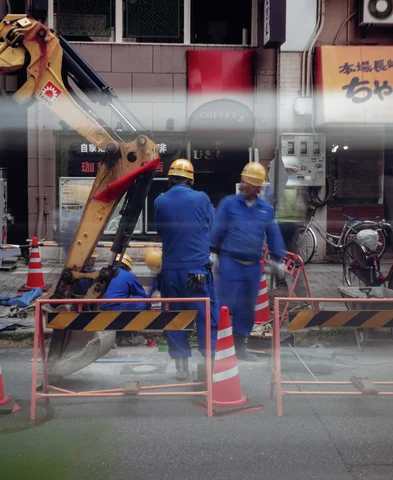 Two workers in blue uniforms and yellow helmets stand beside an excavator behind striped barriers through reflected glass.