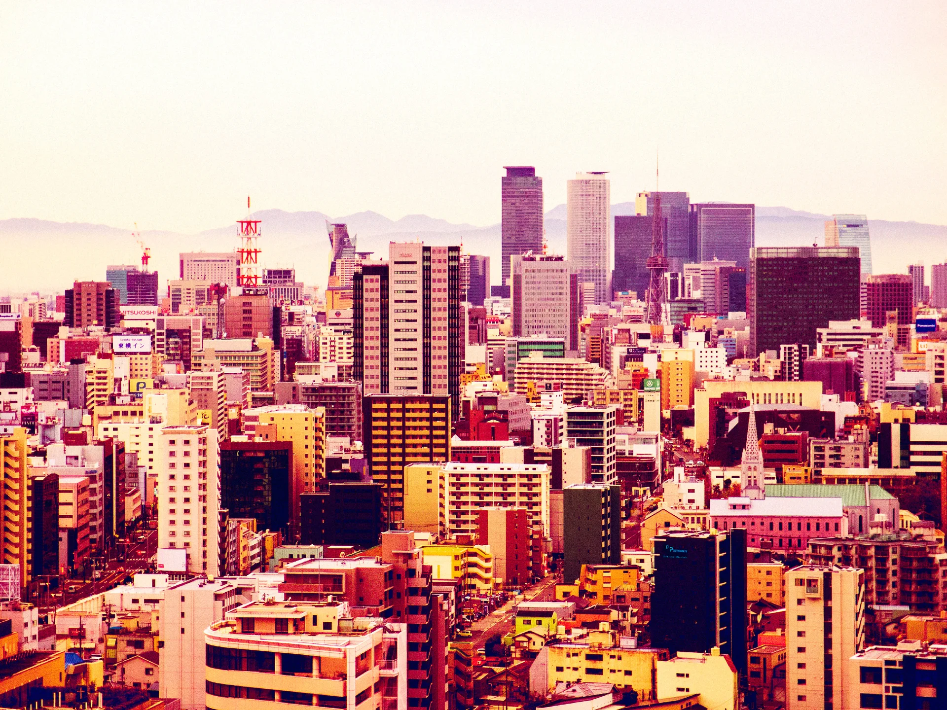 Clusters of pink and orange city buildings rise toward tall towers beneath a pale sky and distant hills.