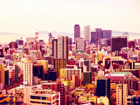 Clusters of pink and orange city buildings rise toward tall towers beneath a pale sky and distant hills.