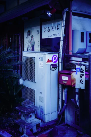 A tall outdoor air-conditioning unit and red mailbox crowd a dark storefront beneath Japanese signs.