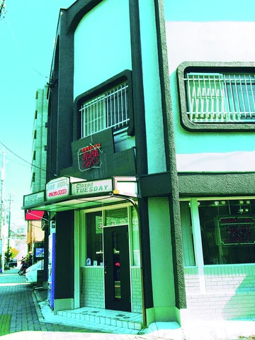 A narrow dark facade surrounds a glass entrance beneath a small awning beside barred windows.