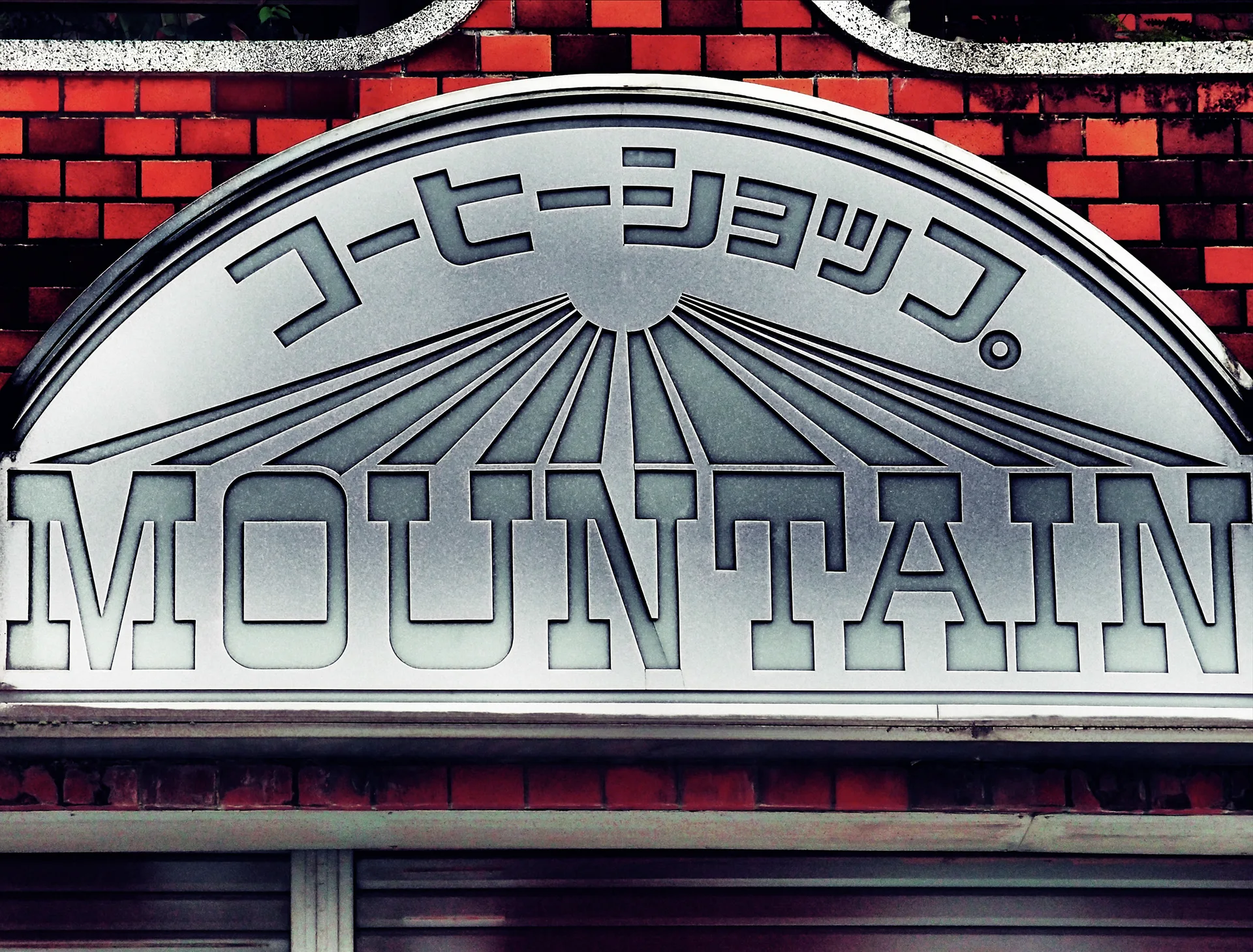An arched silver MOUNTAIN coffee shop sign with radiating lines sits against red brickwork.
